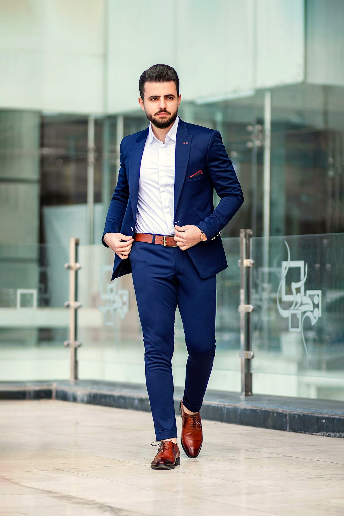 A young man in a navy suit wearing a white Oxford shirt, walking outside a glass office building